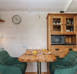A small dining area with a wooden table two green chairs a wooden cabinet with glass doors a toaster oven and plates with croissants and glasses of orange juice at Lottie's Hut in Okehampton