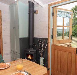 A dining area with a wooden table set with plates of croissants and glasses of orange juice a green chair a wood burning stove by a cabinet and a wooden door open to a view of trees and fields at Lotties Hut in Okehampton