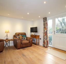 A living room with two leather armchairs a small wooden table a lamp a wall-mounted television a wooden console table with board games floral curtains and a glass door at The Studio at Dan y Garn in Newport Pembrokeshire