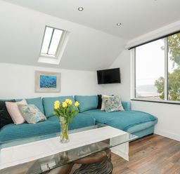 A living room with a blue sofa a glass coffee table with yellow flowers and a window showing trees at Moons Cottage in Fowey