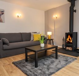 A living room with a gray sofa yellow cushions a black coffee table with flowers a wood burning stove and a lamp at Harran Annex in Peopleton
