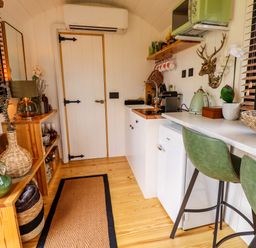A small kitchen with white cabinets green chairs a countertop shelving and various kitchen appliances at Herdwick Luxury Glamping in Nottingham