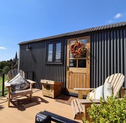 A small outdoor deck with two wooden chairs with cushions a wooden box and a door with a wreath on a black corrugated building at Southdown Luxury Glamping in Nottingham