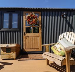 A wooden chair with cushions and a boot box outside a door with a wreath on a corrugated metal building at Southdown Luxury Glamping in Nottingham