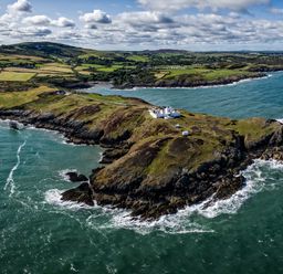 An aerial view of a rocky coastal headland with a lighthouse and buildings surrounded by the sea and fields at East Point Lynas Lighthouse Keeper's Cottage Llaneilian near Amlwch