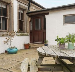 An outdoor patio area with potted plants wooden picnic table and bench in front of building with windows and a wooden door at West Point Lynas Lighthouse Keeper's Cottage Llaneilian near Amlwch