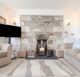 A living room with a stone fireplace and wood stove between two beige sofas with bird patterned cushions at Crab Pot Cottage in Porthleven