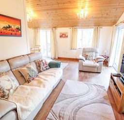 A living room with a leather sofa and armchair with cushions a rug a wooden TV stand and paintings at Kantankye Lodge Thorpe St Peter near Wainfleet All Saints