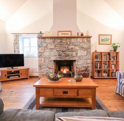 A living room with a stone fireplace a wooden coffee table striped armchairs a television bookshelf and wooden flooring at Rhyd Y Bont Bach in Rhoscolyn