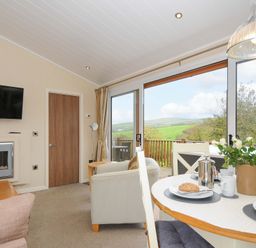 A living room with dining table set with cups and bread and a large window showing green hills at Hideaway Lodge Bossiney near Tintagel