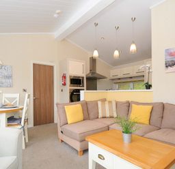 A living room with a brown sectional sofa and yellow cushions a wooden coffee table and a dining table with chairs at Hideaway Lodge in Bossiney near Tintagel