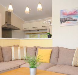 A living room with a beige sofa yellow and striped cushions a wooden coffee table with a plant kitchen area with hanging lights and appliances in the background at Hideaway Lodge Bossiney near Tintagel