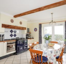 A kitchen with a round dining table and wooden chairs next to a stove and oven with patterned curtains at Bwthyn in Borth-y-Gest near Porthmadog