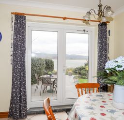 A dining area with table and chairs facing glass doors leading to a patio with table and chairs and a view of water and hills at Bwthyn in Borth-y-Gest near Porthmadog