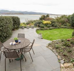 An outdoor patio with a round table and four chairs overlooking a garden and water at Bwthyn in Borth-y-Gest near Porthmadog