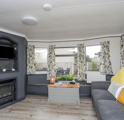 A living room with a gray sofa yellow and white pillows a small table with a lamp books and plants a television above a fireplace and patterned curtains at Rhiwlas Bach in Benllech