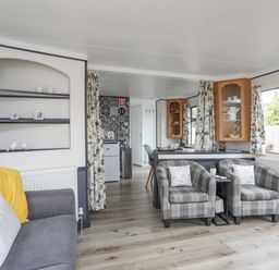 A living room with two plaid armchairs a gray sofa with yellow and white pillows shelves with pictures a small kitchen area with patterned wall tiles and wooden cabinets at Rhiwlas Bach in Benllech