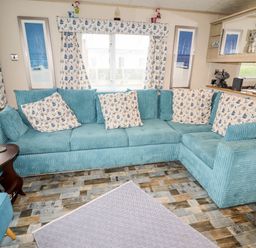 A living room with a blue corner sofa floral curtains and cushions wooden side table and patterned rug at Beachcomber D35 in Towyn