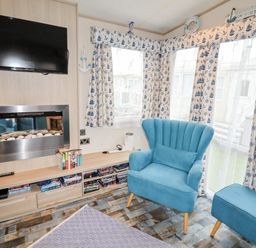 A sitting area with a blue armchair and ottoman near windows with nautical curtains a wall-mounted television and fireplace at Beachcomber D35 in Towyn