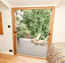 A bedroom with a bed covered in a patterned blanket opening onto a wooden deck with two wooden lounge chairs and green trees outside at The Happy Hut in Sopley near Christchurch