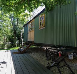 A green metal cabin with wooden door and windows on a gravel area next to a wooden deck with a barbecue grill nearby at The Happy Hut in Sopley near Christchurch