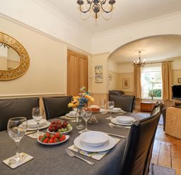 A dining table set with plates glasses and fruit in a room connected to a living area with chairs and a window at Green Gate Cottage in Reydon near Southwold
