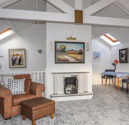 A living room with an armchair and ottoman a fireplace a dining table with two chairs and paintings on the wall at Seaside Sanctuary in Trearddur Bay
