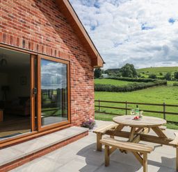 A patio with a wooden picnic table and wine glasses outside a brick house with countryside fields at Awel y Bryn in Rhayader