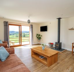 A living room with brown sofas and a wooden coffee table near a wood stove and sliding glass doors at Awel y Bryn in Rhayader