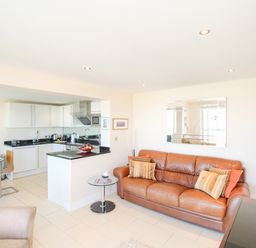 A living room with a brown leather sofa and a kitchen area with white cabinets at Sunset View in Castletown Portland