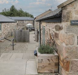 An outdoor stone walkway between buildings with a wooden fence and patio furniture at The Coach House in Bamburgh