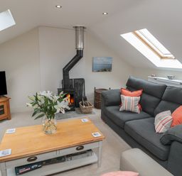 A living room with a gray sofa and wooden coffee table with flowers by a wood burning stove at The Coach House in Bamburgh