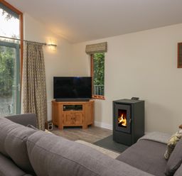 A living room with a gray sofa a wood burning stove a wooden TV stand with a television and windows showing greenery at Bay Tree Lodge in Cartmel