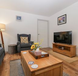 A living room with a wooden coffee table with a tray of flowers a grey armchair a floor lamp a television on a wooden stand a framed picture on the wall at Ty Glas in Ogmore-by-Sea near St Brides Major