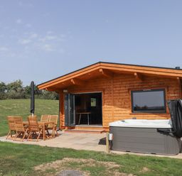 A wooden cabin with an outdoor dining table and chairs and a hot tub on a patio at Foxglove Lodge in Hittisleigh
