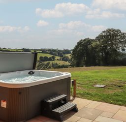 An outdoor hot tub on a tiled patio with steps and a view of green fields and trees at Bluebell Lodge in Hittisleigh