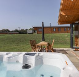 An outdoor patio with a hot tub and wooden table with chairs near a wooden cabin at Bluebell Lodge in Hittisleigh