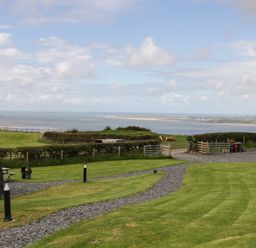 A grassy garden with a gravel path and a wooden picnic table overlooking a coastal landscape with water and hills at Orchard in Dalton-In-Furness