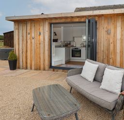 An outdoor seating area with a wicker sofa and table in front of a wooden cabin with open doors showing a kitchen at Hillside Hut Radbourne near Ashbourne