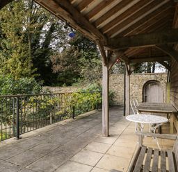 An outdoor covered patio with wooden bench wooden table white metal chairs and a stone wall with an arched wooden door at The Barn at Westhall Cottage Fulbrook near Burford