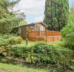 A wooden cabin with a fenced porch near a small stream surrounded by trees and bushes at Willows Lodge White Cross Bay near Windermere