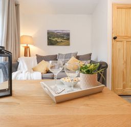 A living room with a wooden table holding a tray with two empty wine glasses a bowl of popcorn and a potted plant a gray sofa with cushions and a white throw a floor lamp and a wooden door at The Wonky Burrow in Bowness-On-Windermere