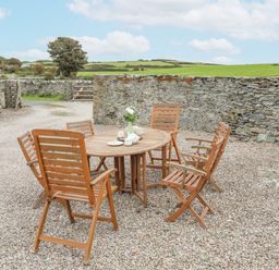 An outdoor seating area with a round wooden table and six wooden chairs on gravel surrounded by stone walls at Caerdegog Ganol in Cemlyn near Cemaes Bay