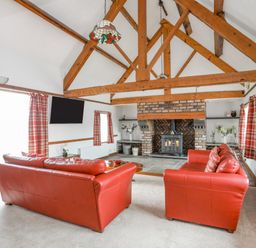 A living room with red leather sofas, a brick fireplace, exposed wooden beams and red plaid curtains at Caerdegog Ganol in Cemlyn near Cemaes Bay