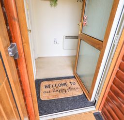 An open wooden front door revealing a welcome mat with text and an interior hallway with a heater on the wall at Our Happy Place Lakeside 16 Borwick near Carnforth