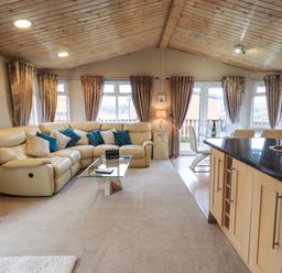 A living room with a beige sectional sofa and blue cushions next to a glass coffee table a dining table with chairs and a kitchen island with a sink at Our Happy Place Lakeside 16 Borwick near Carnforth
