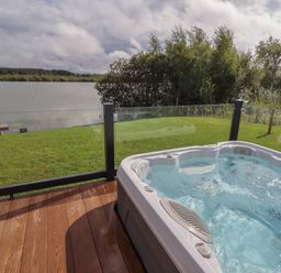 An outdoor hot tub on a wooden deck overlooking a grassy area with a lake and trees at 40 Delamere Point in Delamere