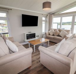 A living room with three beige sofas, a wooden coffee table, a wall-mounted TV, and a fireplace with large windows overlooking a lake at 40 Delamere Point in Delamere