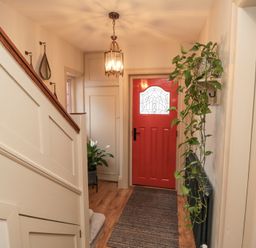 A hallway with a red front door a hanging light fixture plants on the floor and wall and a staircase with white paneling