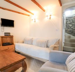 A living room with a wooden table sofa and armchair a wooden cabinet with a television on the wall and stairs with a stone wall at Aunt Dora's in Port Isaac